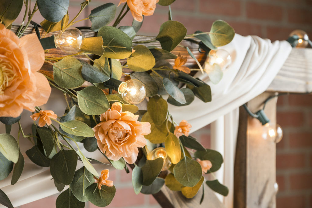 A garland of bright paper flowers wrapped around string lights on a patio frame. 
