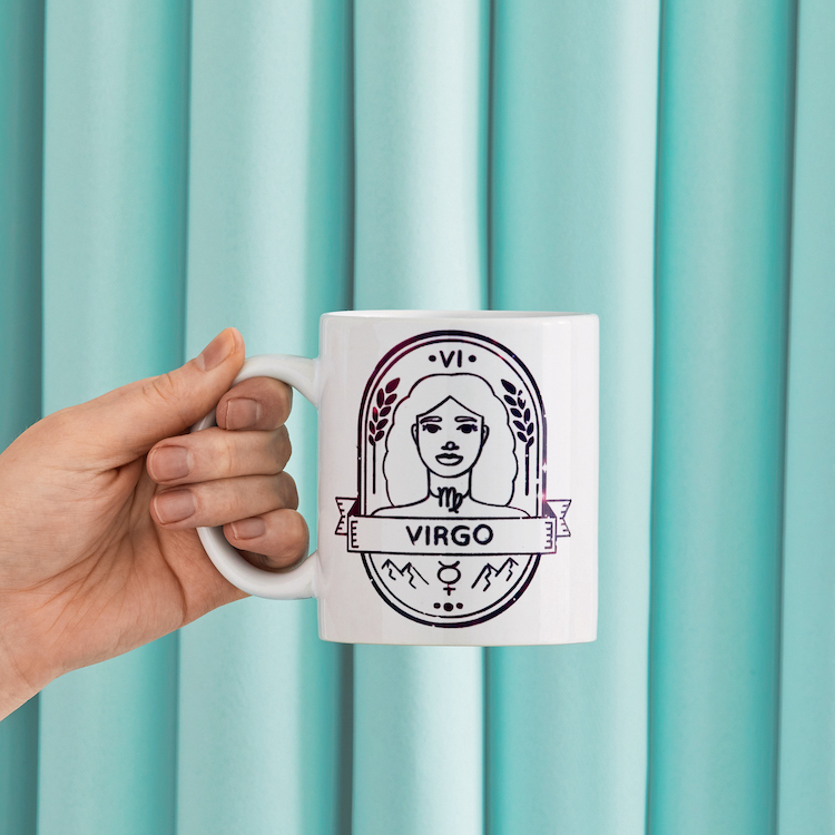 Hand holding coffee mug with "Virgo" Zodiac symbol in front of blue background