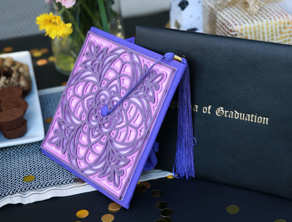 Purple graduation cap design with an ornate layered design, sitting next to a diploma on a black table cloth.