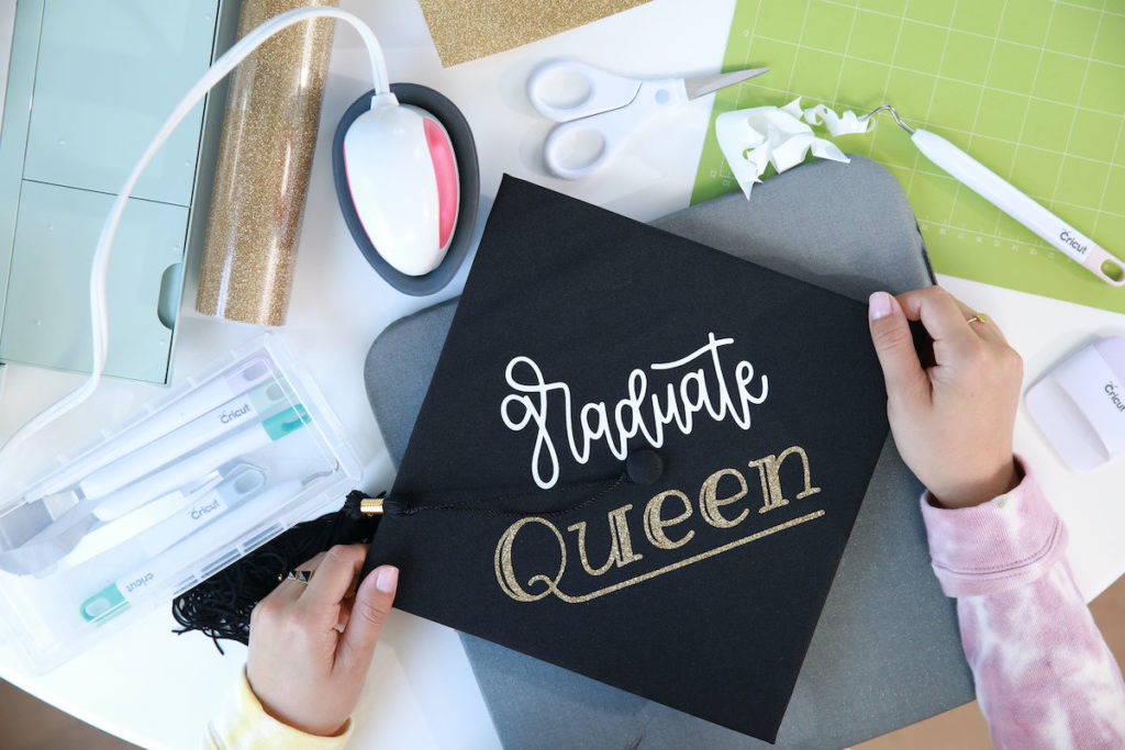 Hands crafting a black graduation cap design that reads "graduate queen" on top with cursive font