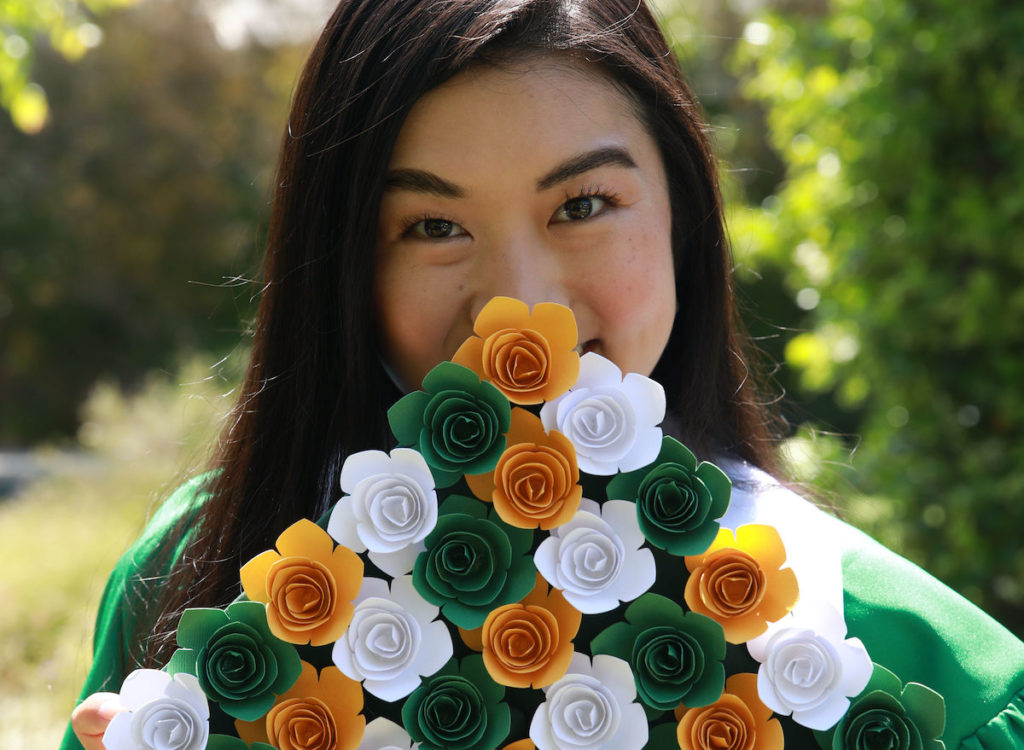 A girl holds up a grad cap in front of her face– the graduation cap design is covered in yellow, green, and white paper flowers.