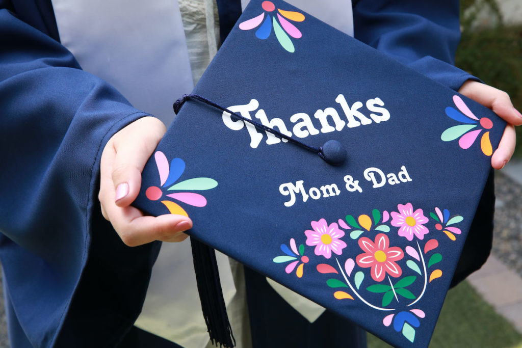 A girl holds out a grad cap that reads "Thanks mom and dad". The graduation cap design is blue and embellished with colorful vinyl flowers.