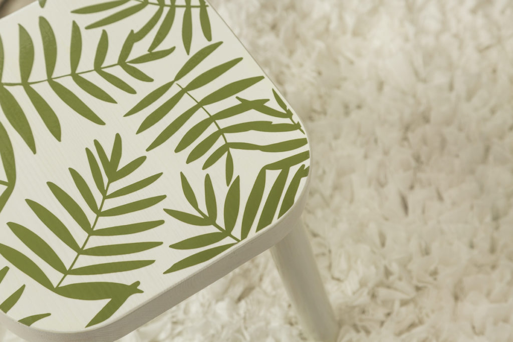 A trendy stool with palm leaf vinyl decals sits on a white fluffy rug.