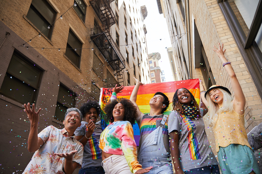 A group of diverse young millennials wearing colorful clothing hold a pride flag as they march through an alleyway