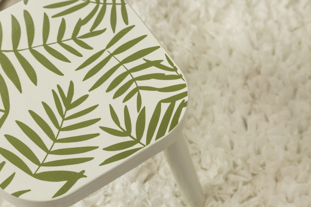 A trendy stool with palm leaf vinyl decals sits on a white fluffy rug.  
