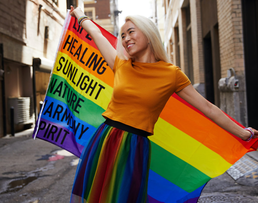 A woman with bright blonde hair and a rainbow skirt holds up a Pride flag in an alleyway