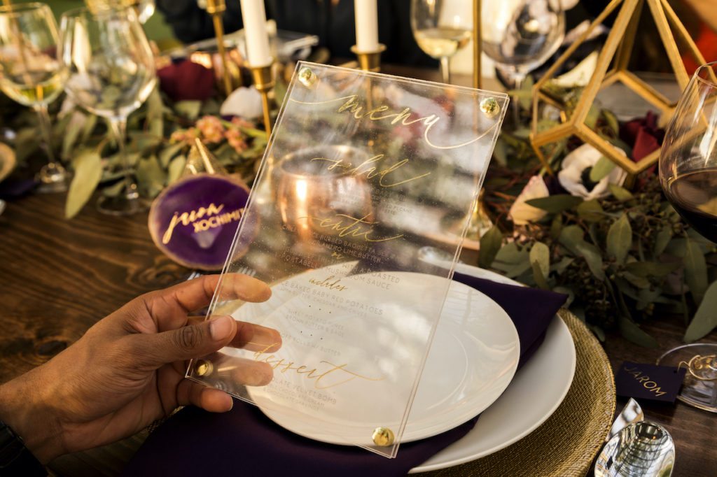 A hand holds a clear, acrylic menu gilded with golden script that lists the dinner menu items. 