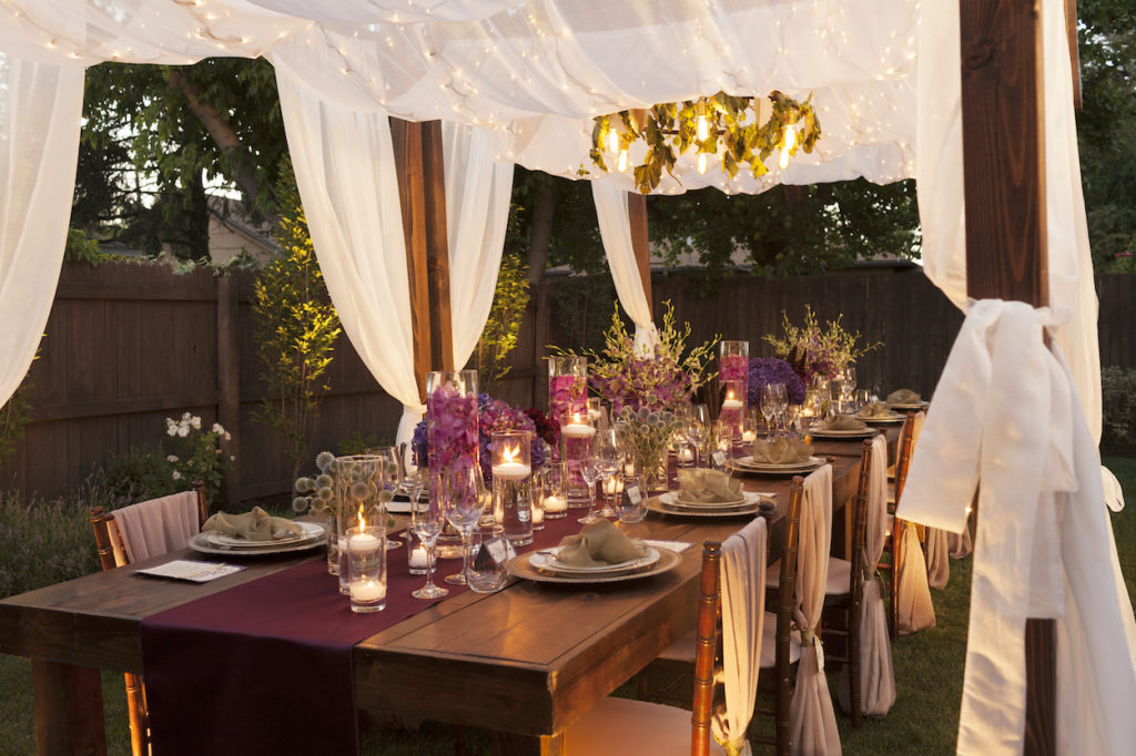 An outdoor table glows under candlelight, covered by a linen wrapped gazebo decked out in flowers and greenery.