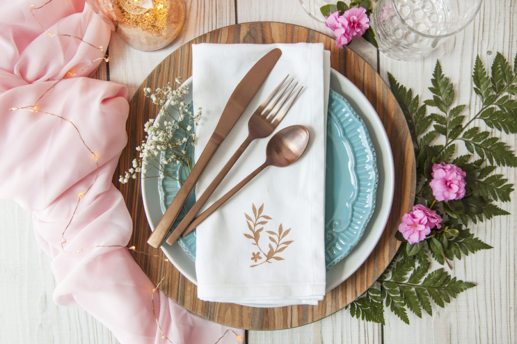 A wooden and floral table setting sits atop a rustic table, complimented by copper cutlery and soft pink cotton table accents. 