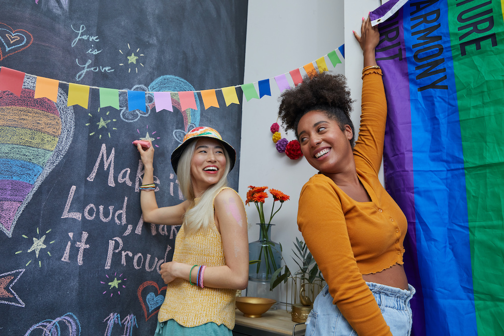 Two people decorate their apartment walls with rainbow colored decor to celebrate Pride month