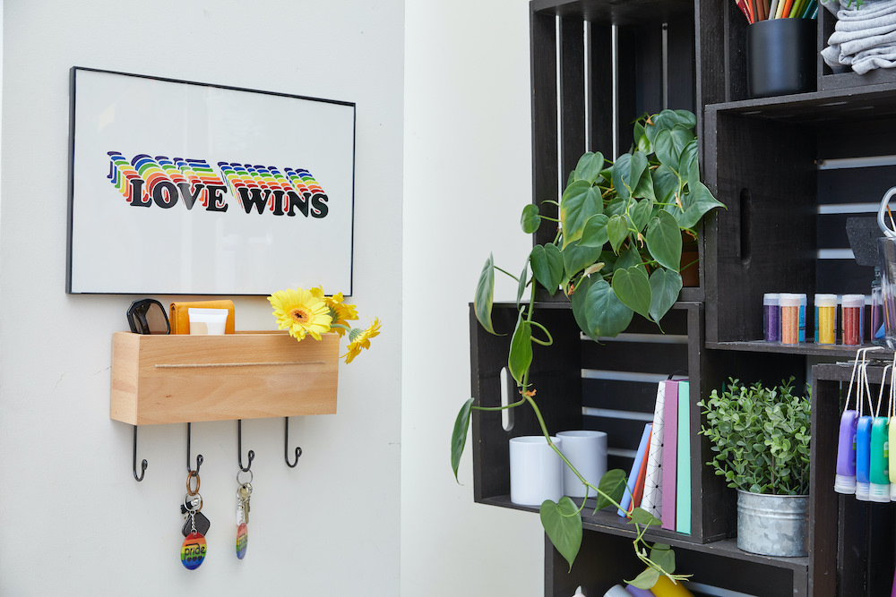 A graphic rainbow pop art of the phrase "love wins" sits on a white wall next to a black crafting shelf and key holder. 