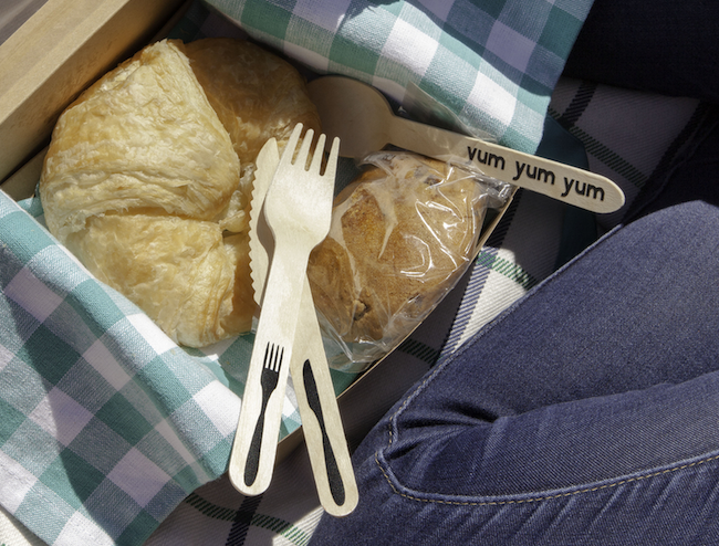 Wooden cutlery sits inside a cardboard picnic box, next to a croissant and muffin
