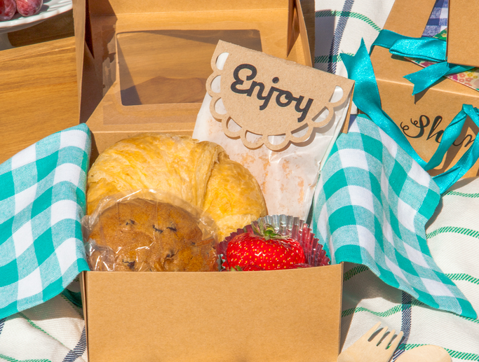 Fruits and pastries sit inside a cardboard picnic box with a blue gingham napkin