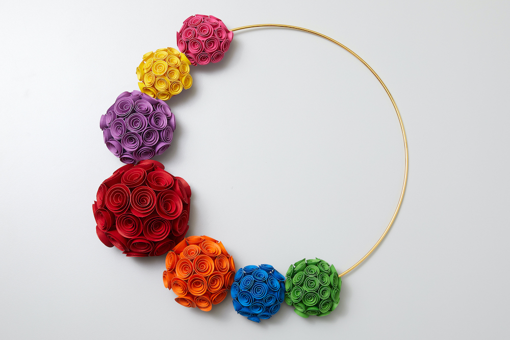 A rainbow colored wreath made of rolled paper roses hangs along a white wall. 