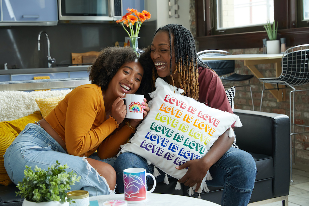 Two people sit together laughing on a couch, holding a white pillow that reads "love is love" in various colors. 
