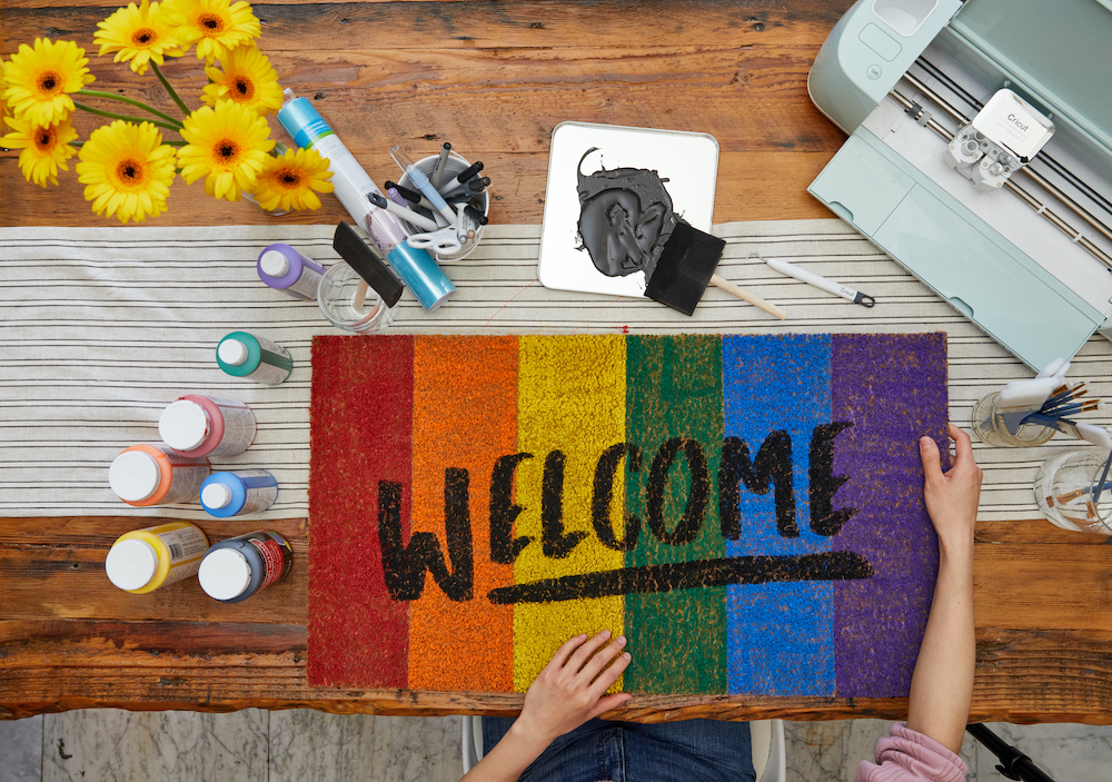A pair of hands holds a Pride Party welcome mat that reflect the rainbow colors of the gay Pride flag.
