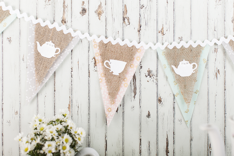 Outdoor tea party decor idea.  A burlap and pastel colored banner with tea party motifs sits on a farmhouse wall
