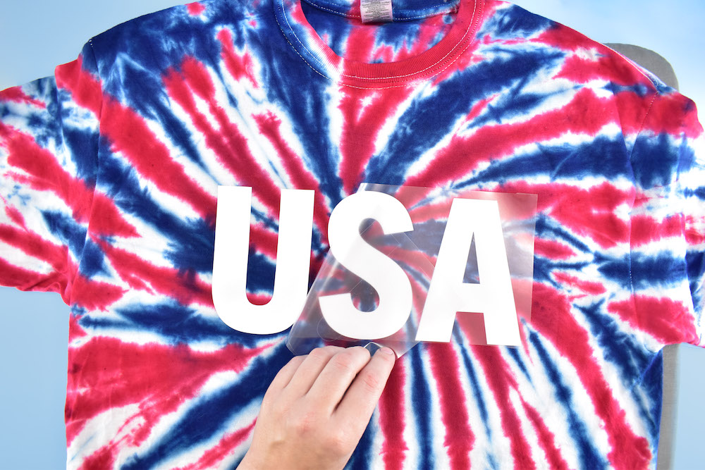 Plastic is peeled from a vinyl "USA" design applied to a red, white, and blue tie dyed tee