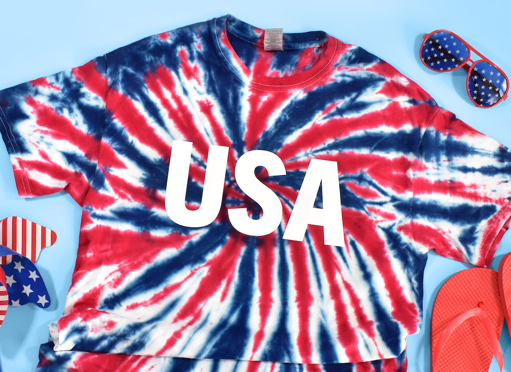 A red, white, and blue tie-dyed USA t-shirt for the 4th of July is surrounded by patriotic flip flops and sunglasses