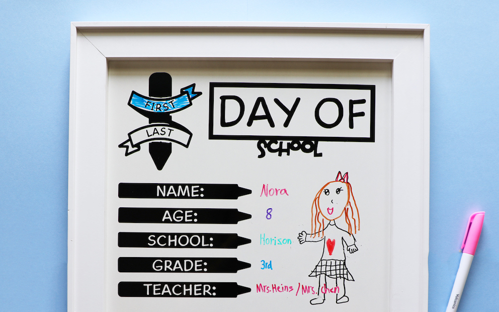 A back-to-school photo shoot white board sits on a blue background; it has child illustrations and handwriting