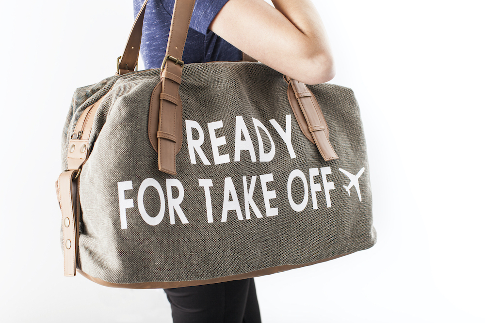 A personalized, brown carry-on canvas duffel has a design that reads "ready for takeoff" in white iron-on vinyl
