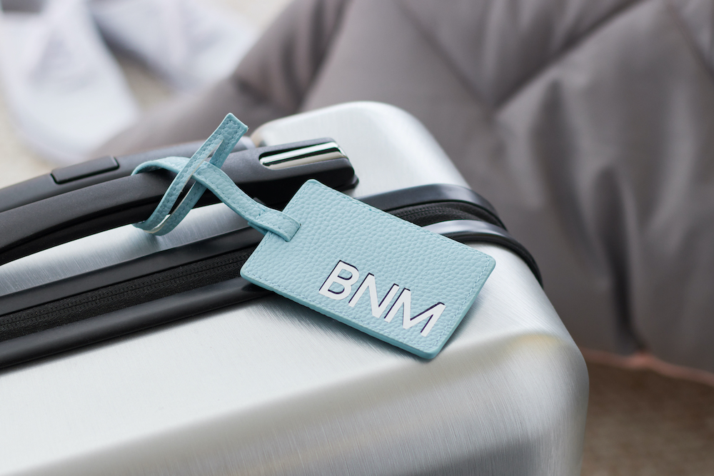 A blue monogrammed luggage tag sits on a silver rolling suitcase