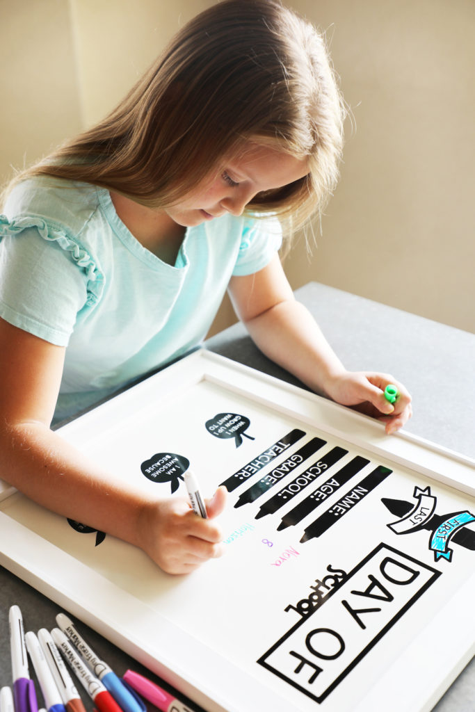 A young girl decorates a back-to-school photo shoot white board prop with dry erase markers